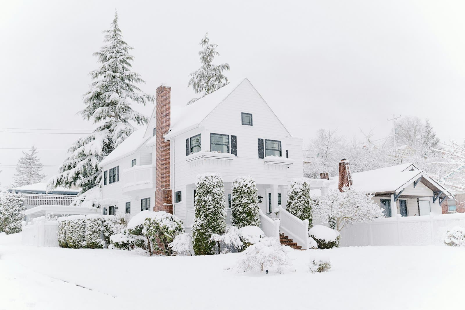 A white two-story house and snow-covered hedges after a heavy snowfall, with power lines and a utility pole along the street.