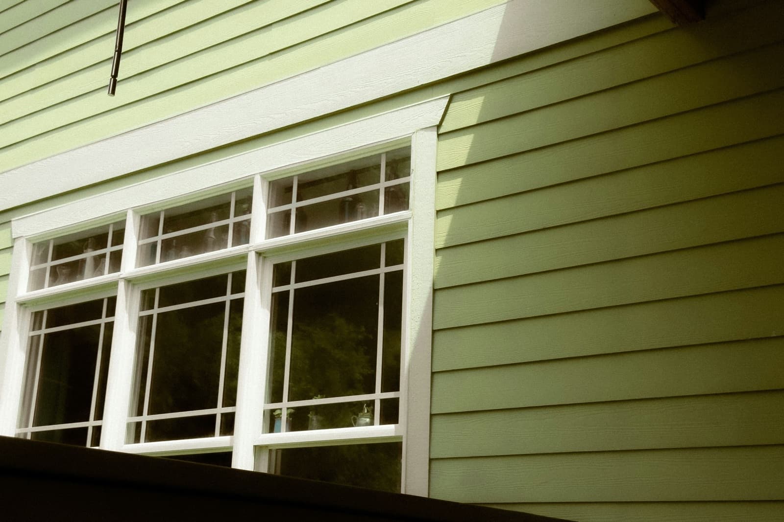 Three white double-hung windows with grid panes, framed in white trim, set into pale green lap siding on the side of a house.