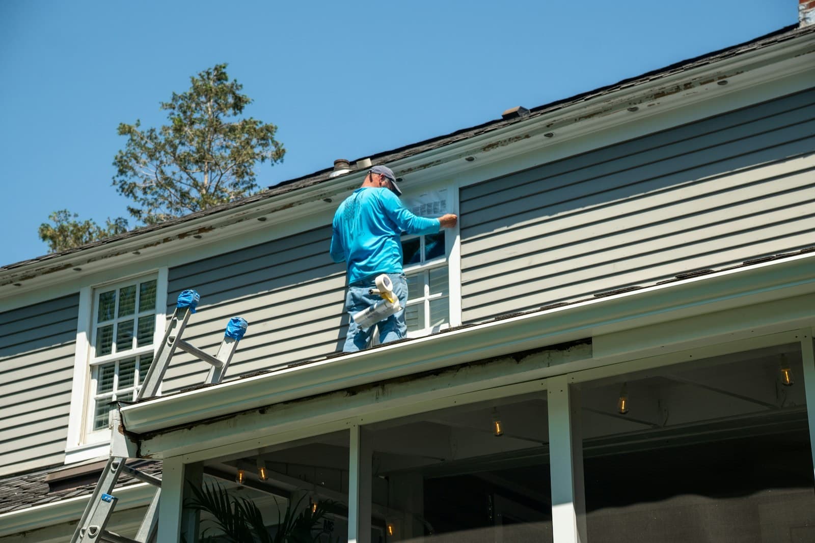 A worker in a blue shirt and cap, seen from behind, holds a printed sheet against an upstairs window on a gray-sided house; a ladder leans against the wall to his left.